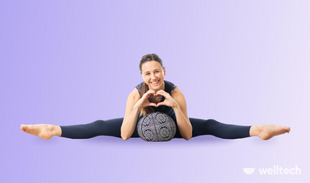 a woman is sitting on the floor with her legs wide in a split, lying on a yoga bolster, with her hands in a heart shape_yoga poses with a bolster