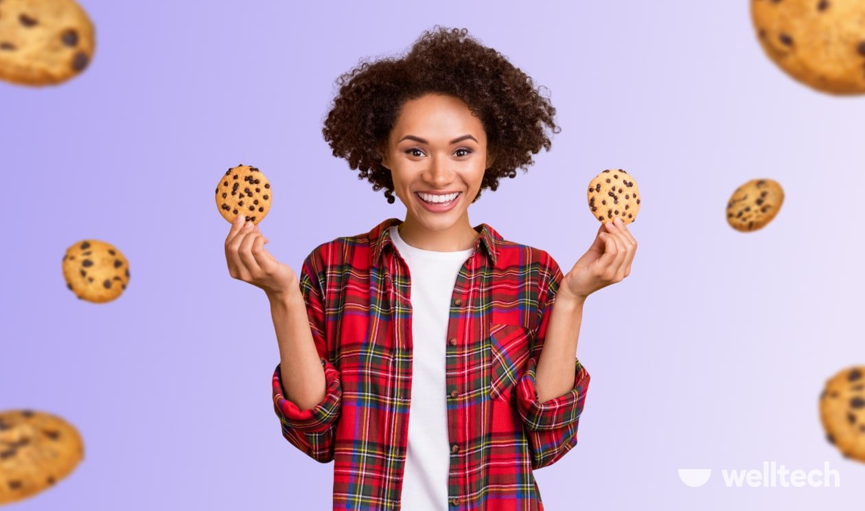 a woman with short curly hair is looking in the camera, smiling, holding cookies in both hands, keto cheat day